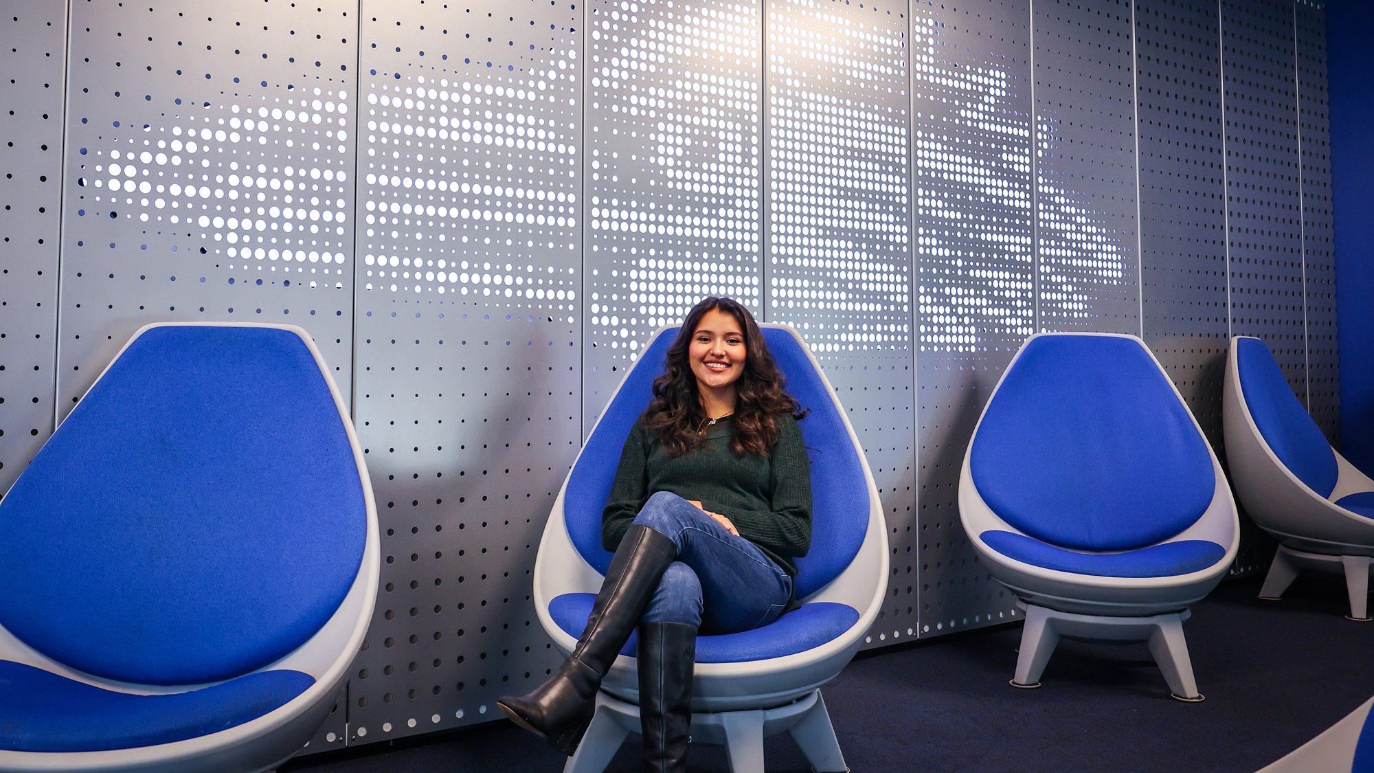 Aerospace engineering technology major Evelyn Montes Gonzalez sitting in front of the white Roadrunner birdhead in the JSSB student lounge in a blue chair