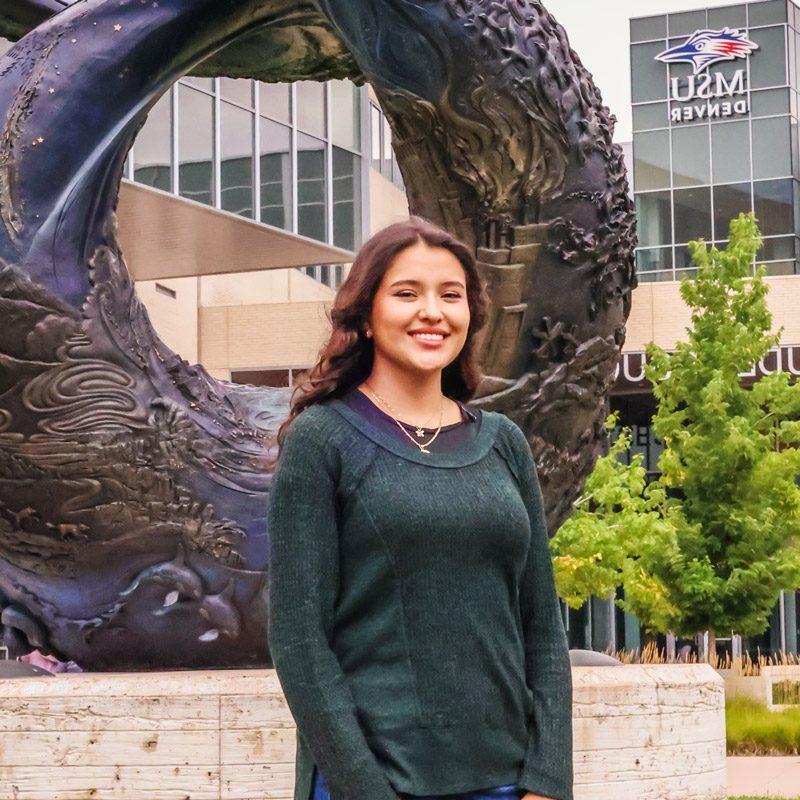 Aerospace engineering technology major Evelyn Montes Gonzalez standing in front of the One World, One Water statue in front of JSSB with the MSU Denver sign in the background