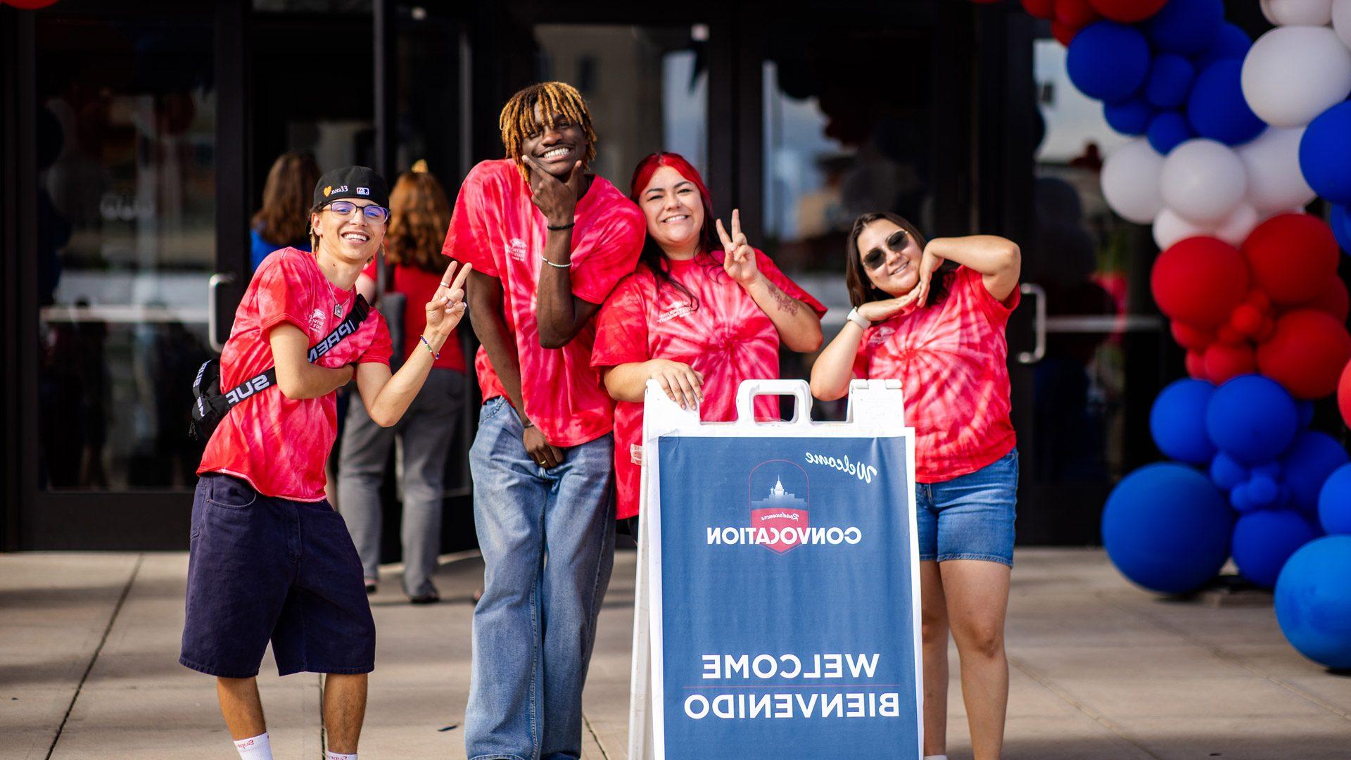 Four students wearing red tie-dye MSU Denver shirts smiling and standing in front of a Welcome/Bienvenido sign at Fall 2024 Convocation.